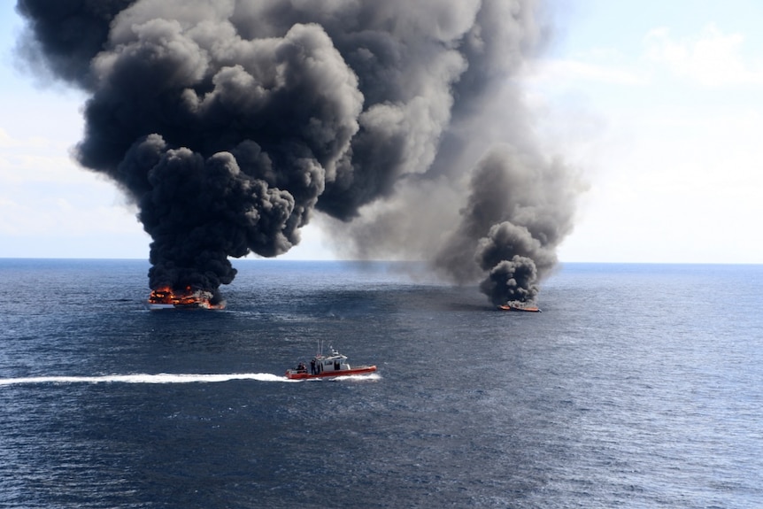 A US Coast Guard boat sails past two vessels on fire in the ocean, billowing out smoke into the sky