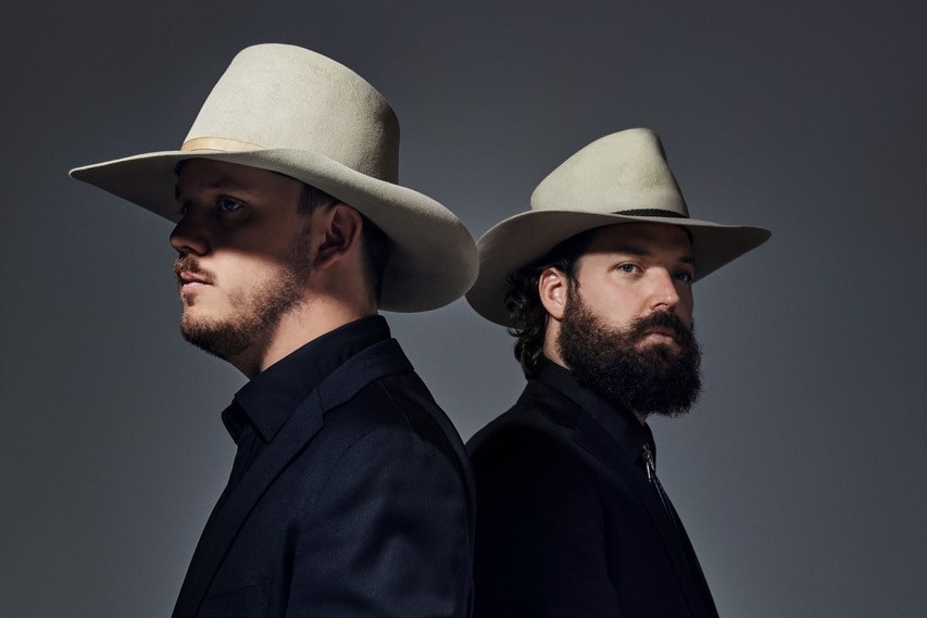 Two men wearing Akubras, in all black with facial hair standing side by side