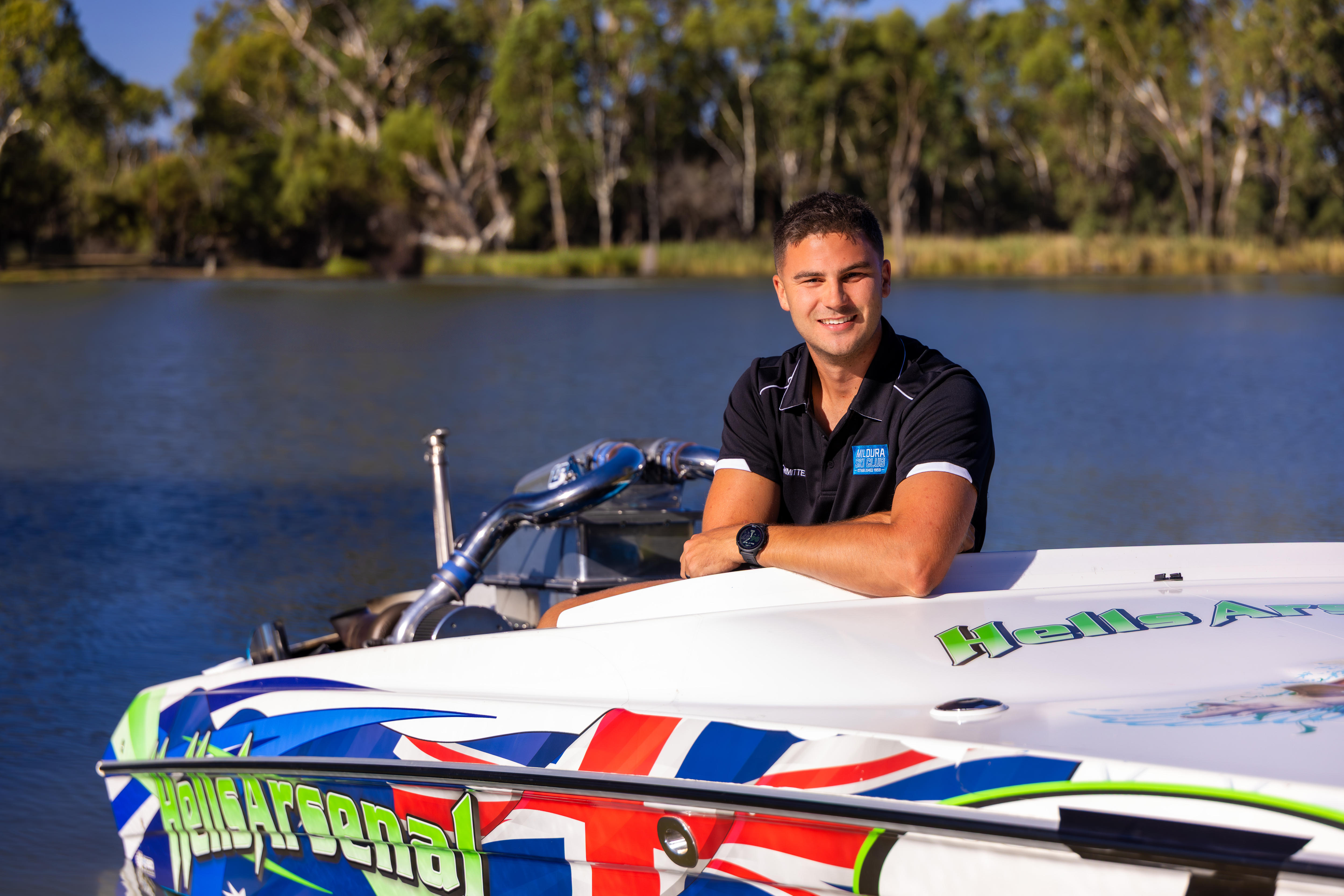 Un hombre con un polo negro cerca de un río, apoyado en un bote de esquí con “Hells Arsenal” escrito en letras verdes.