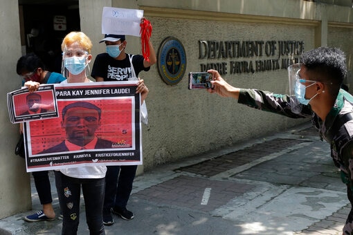 A person holds red signs out the front of a court. A man with a smart phone wearing a face shield is taking a photo of them.