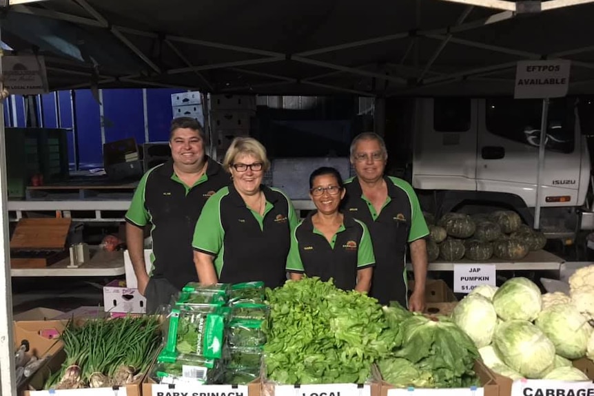four people stand smiling at the camera, in front of a row of green produce