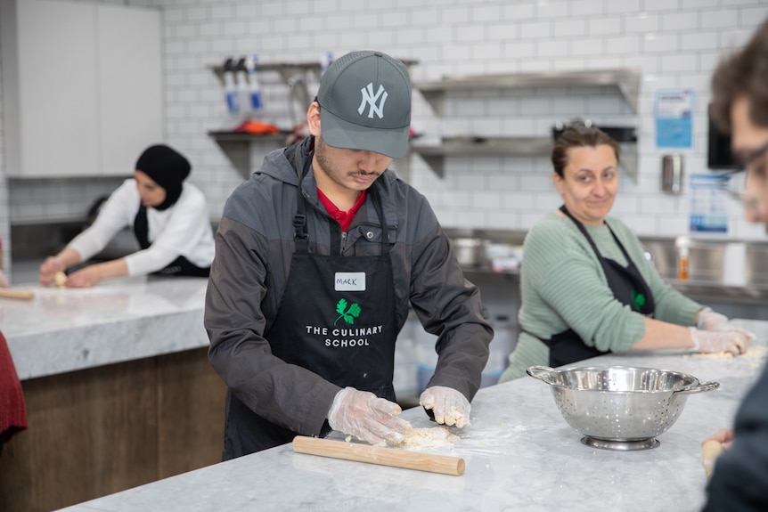 A man in a kitchen, looking down at his hands as he kneads dough. A woman working on the same bench smiles at him.