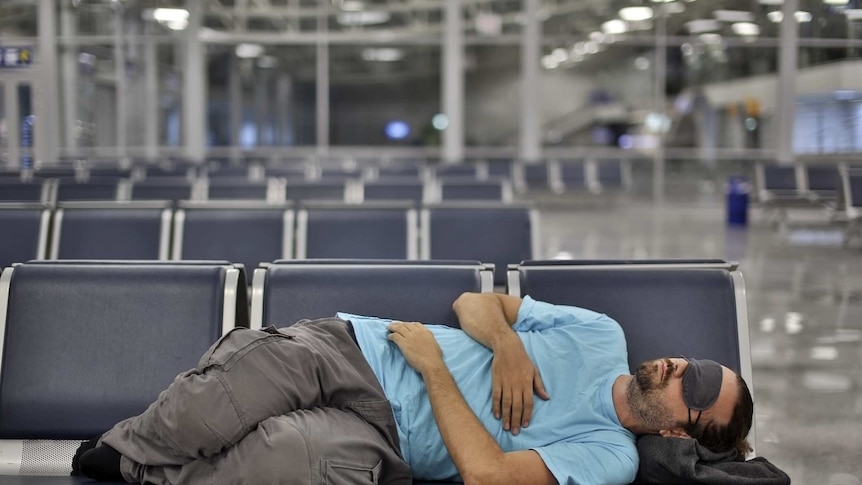 A man wearing an eye mask asleep across several seats at an airport