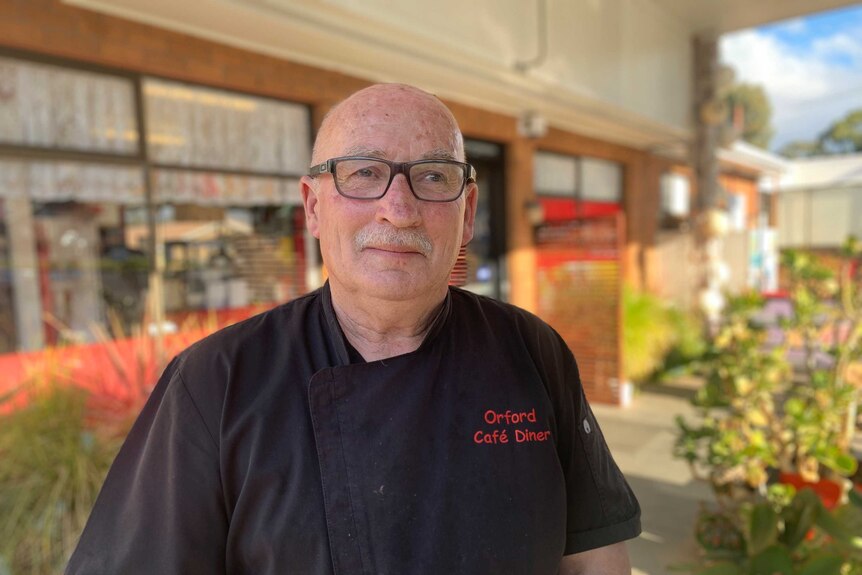 A bald man with a grey moustache, wearing glasses and a black top stands in front of a cafe.