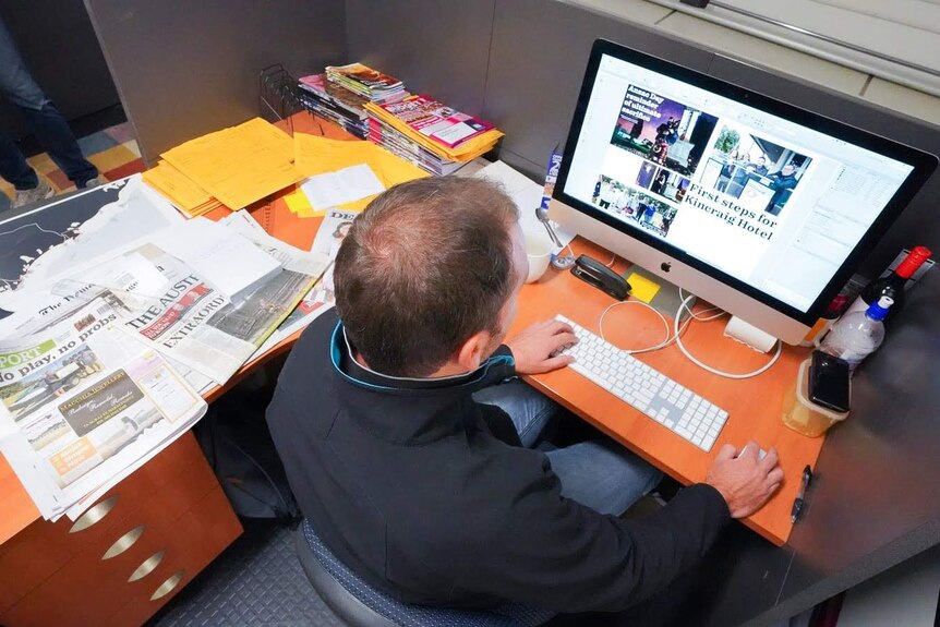 A man sits at a computer working on a newspaper layout with newspapers strewn across his desk.