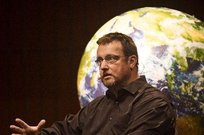 Man speaks at an event, in front of a slide of the globe