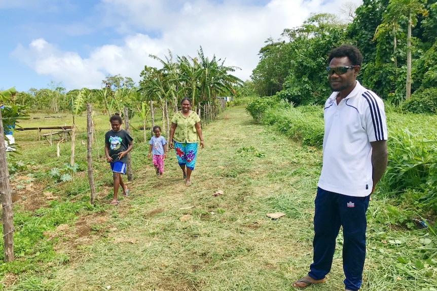 John Bakoa stands near a grassy path next to an empty, tropical paddock. Two children and a woman are walking down the path.