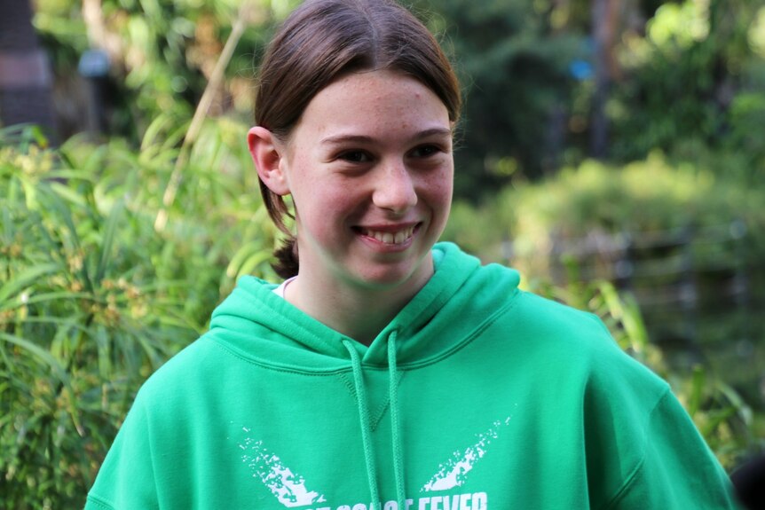 Abagail smiles, wearing a green hooded jumper. There are plants behind her.