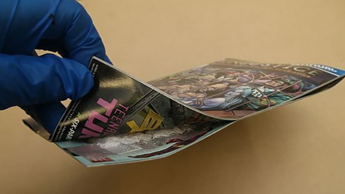 A gloved hand lifts up the corner of a comic book to reveal drugs hidden inside