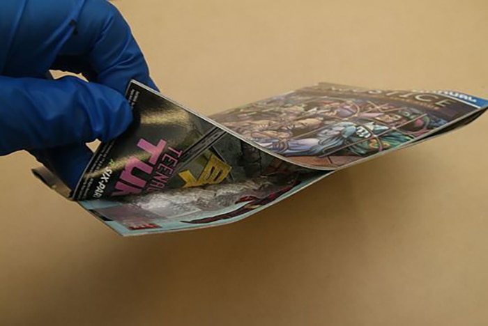 A gloved hand lifts up the corner of a comic book to reveal drugs hidden inside