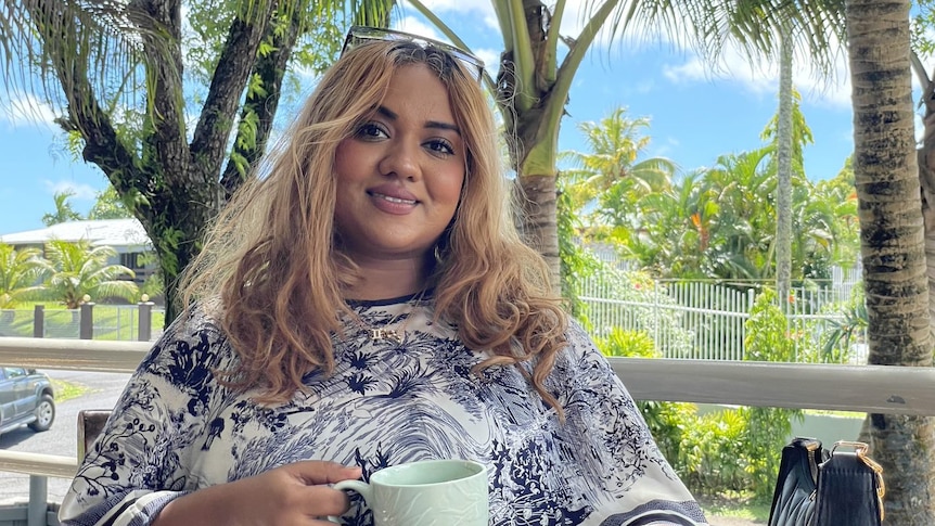 A fijian woman with long bonde hair smiles outside on a sunny day holding a coffee 