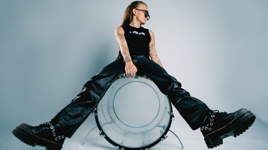 G Flip sits with legs spread on a clear kick drum. They're wearing black leather pants, chunky shoes, and a black sleeveless top