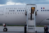 Emmanuel Macron, wearing a black suit, walks up the steps to his Presidential plane while waiving at someone in the distance