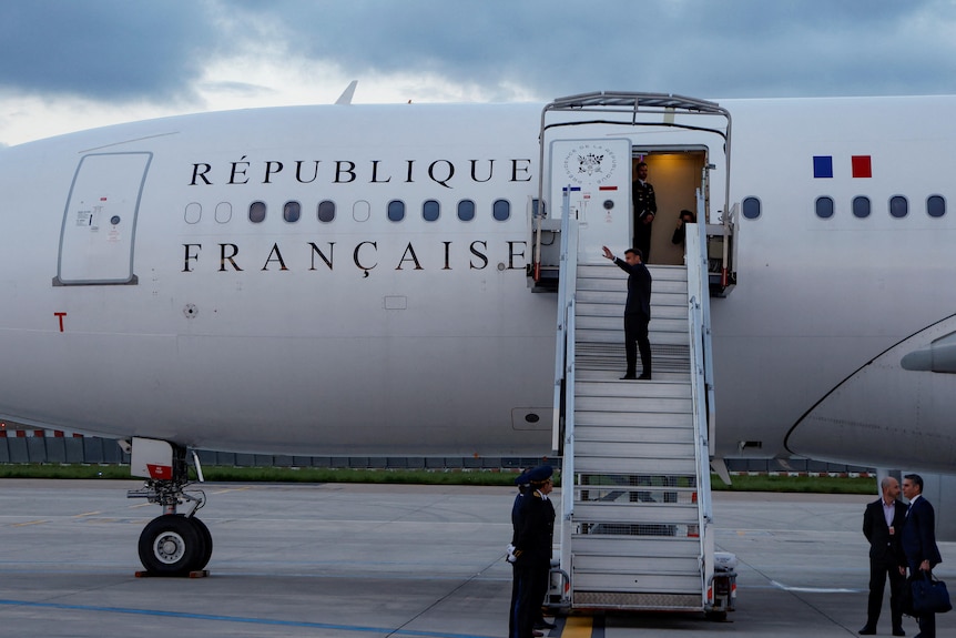 Emmanuel Macron, wearing a black suit, walks up the steps to his Presidential plane while waiving at someone in the distance