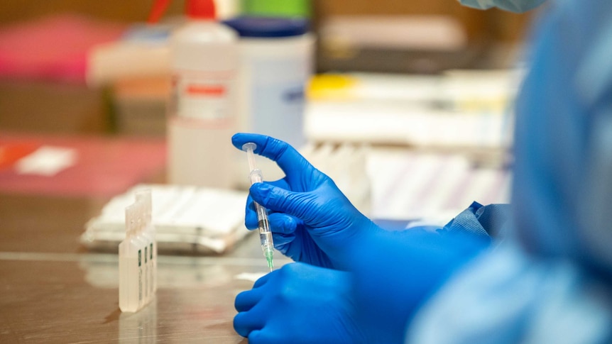 A health worker wears blue latex gloves and draws some coronavirus vaccine from a vial with a syringe. A health worker wears blue latex gloves and draws some coronavirus vaccine from a vial with a syringe.