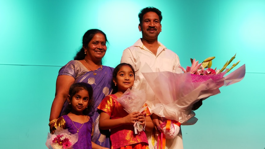 Four people, a mum, dad and two young daughters, stand on a stage holding flowers
