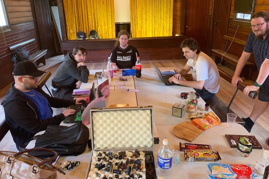 Five males  sit at a table in a historical rural hall, ready to play video games. There's junk food and lap tops on table.