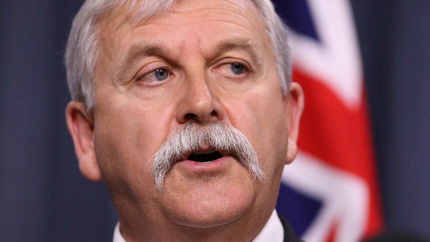 A man with grey hair and a moustache at a press conference in front of an Australian flag.