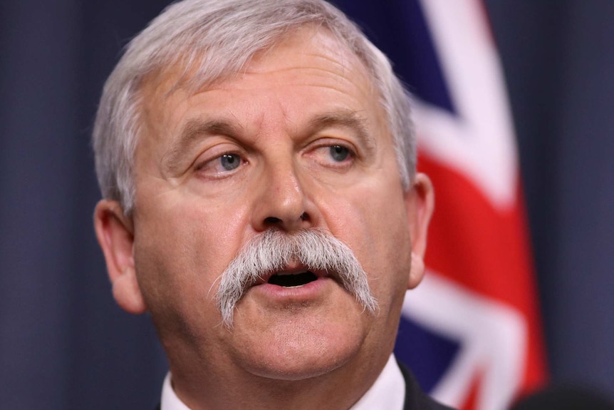 Ein Mann mit grauen Haaren und Schnurrbart bei einer Pressekonferenz vor einer australischen Flagge.