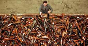 A man lifts a gun into a large metal container containing hundreds of guns.