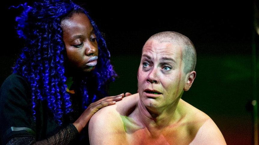 A woman in black lays her hands on the back of a naked woman with a shaved head.