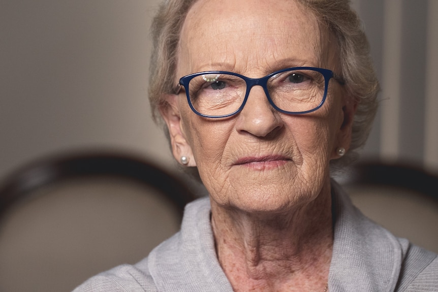 An older woman wearing glasses looks at the camera.
