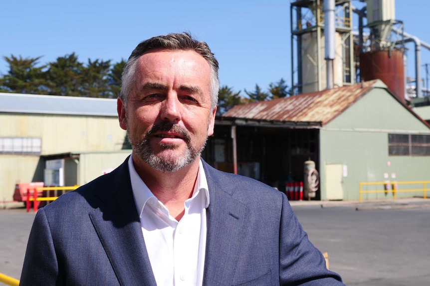 A middle aged man with a salt and pepper beard and hair looking at the camera. He's standing in a sawmill