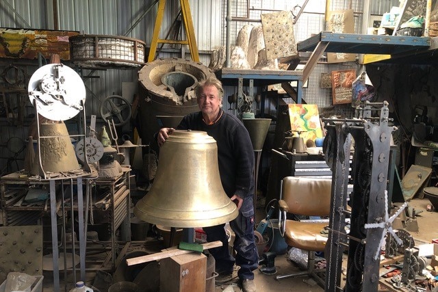a man in a messy workshop area standing behind a large, gold coloured bell he made himself.