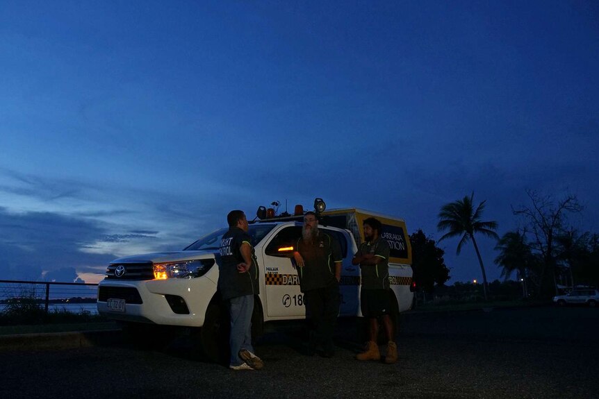 A photo showing men standing in front of a night patrol car at dusk.