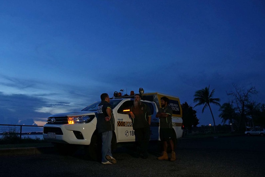 A photo showing men standing in front of a night patrol car at dusk.