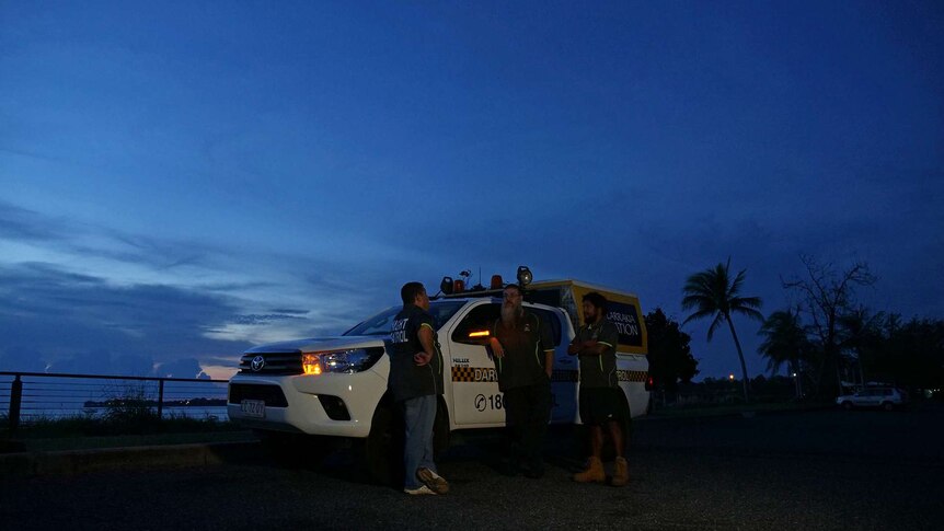 A photo showing men standing in front of a night patrol car at dusk.