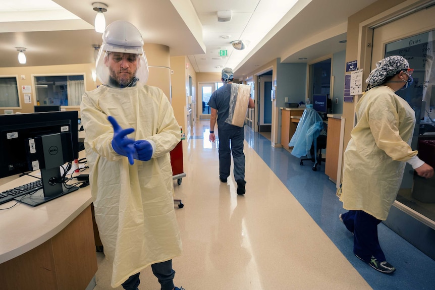 A man in full PPE standing in a hospital ward snaps on blue gloves