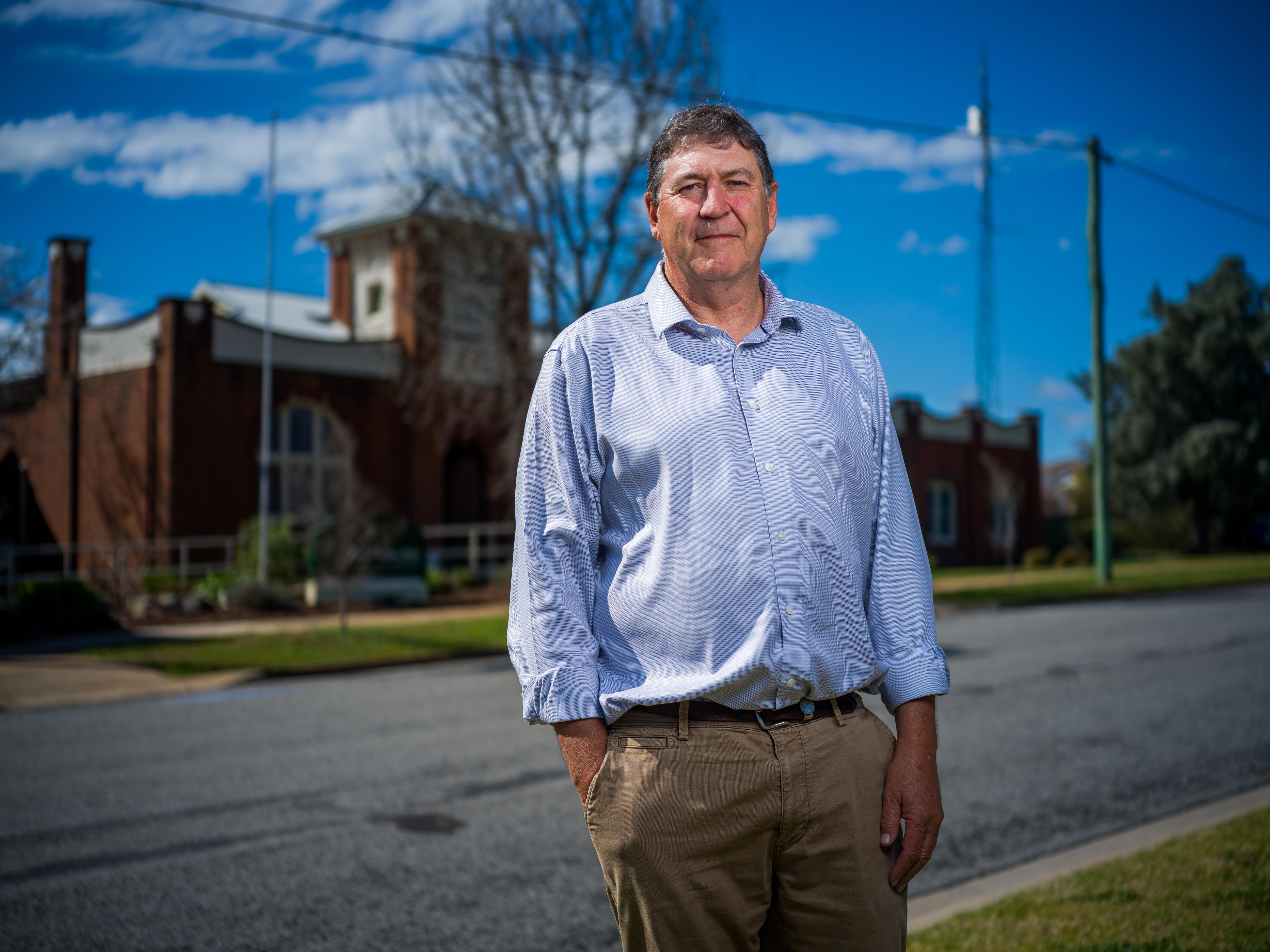 A man looking serious in a blue shirt stands across the road of the council building which is in background