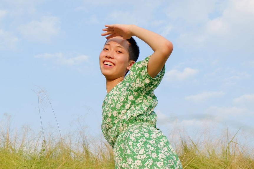 A woman in a green dress with a buzzcut looks back at the camera in the middle of a grassy field