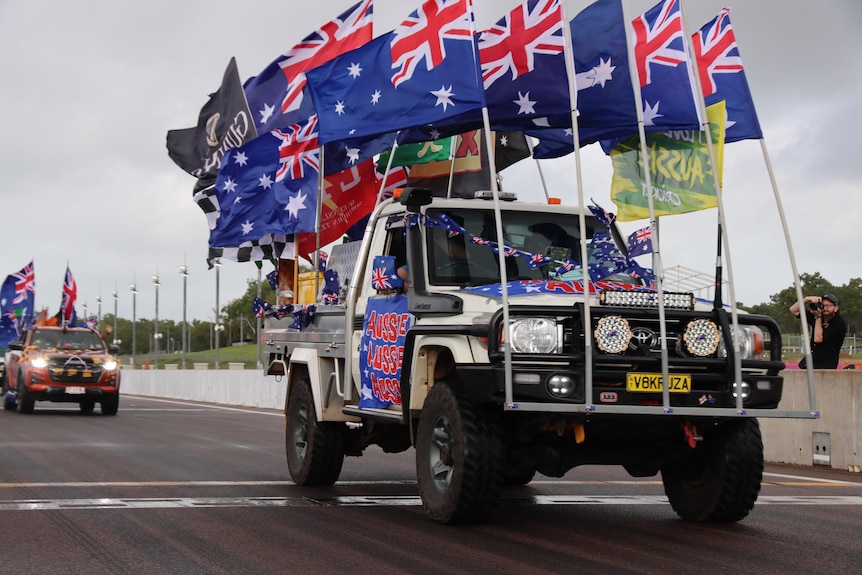 A ute drives along a road flying several Australian and other flags, a banner on the side says "Aussie Aussie Aussie"