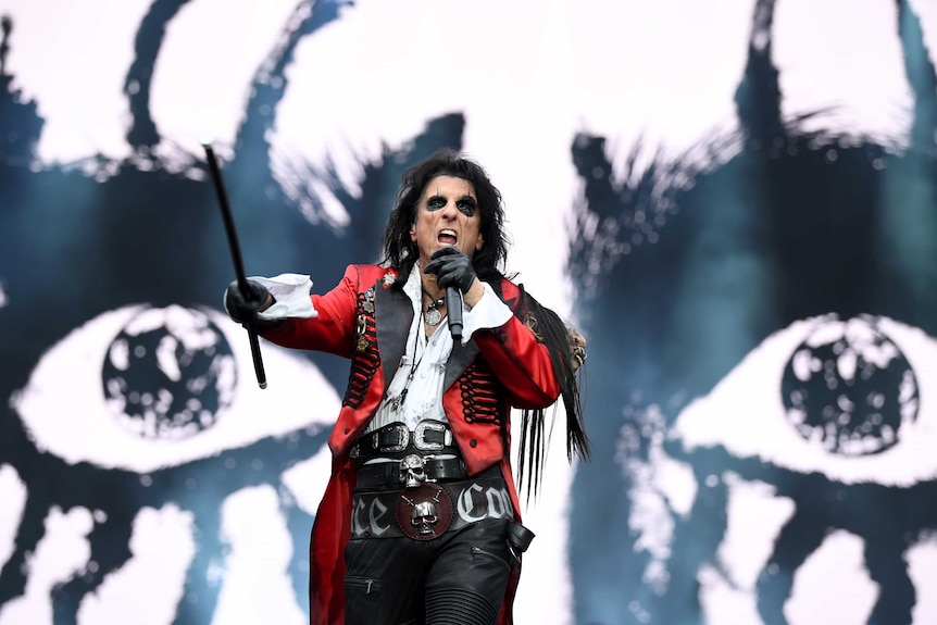 Alice Cooper waving a cane in front of a giant screen with a black and white face.