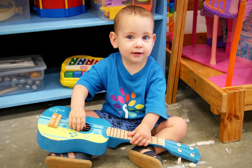 A baby clutches a toy guitar in front of a shelf of toys 