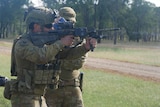 A soldier with a gun firing, a trainer behind him