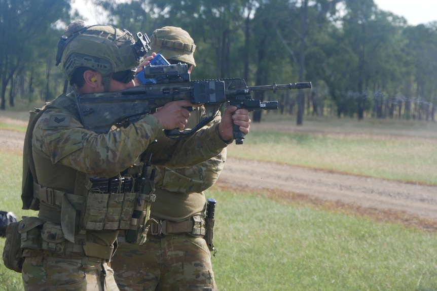 A soldier with a gun firing, a trainer behind him