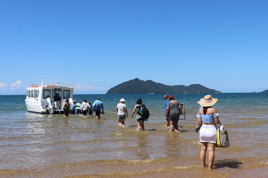 A group of people wade into the water at a beach, some getting onto a boat, with Dunk Island in the background