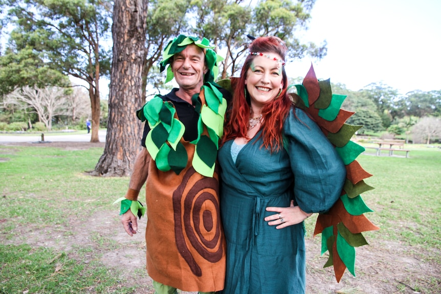 A man dressed up as a tree and a woman dressed up as a fairy stand in a park.