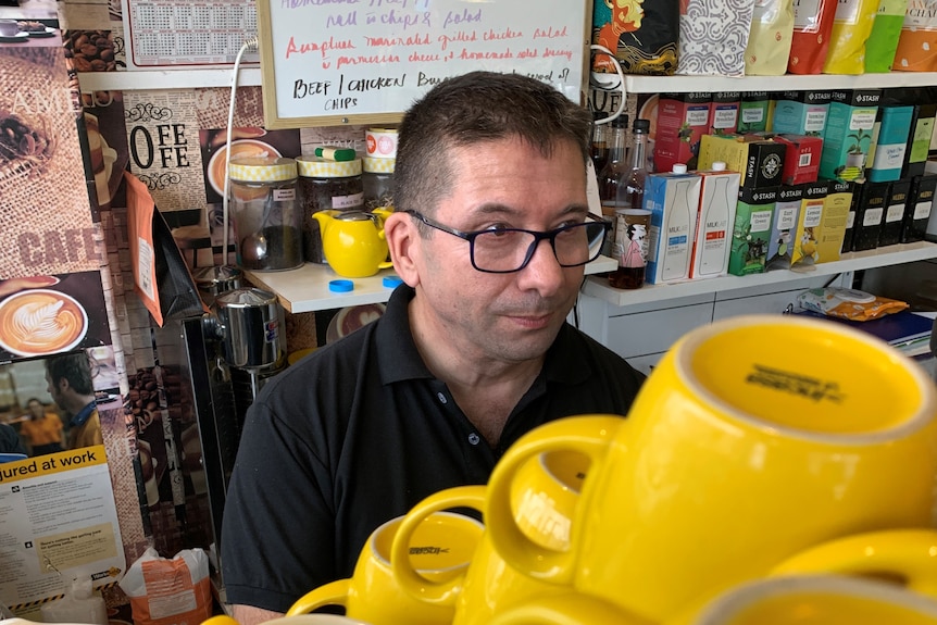 A man with a black polo shirt makes coffee, yellow coffee mugs are stacked in the foreground.