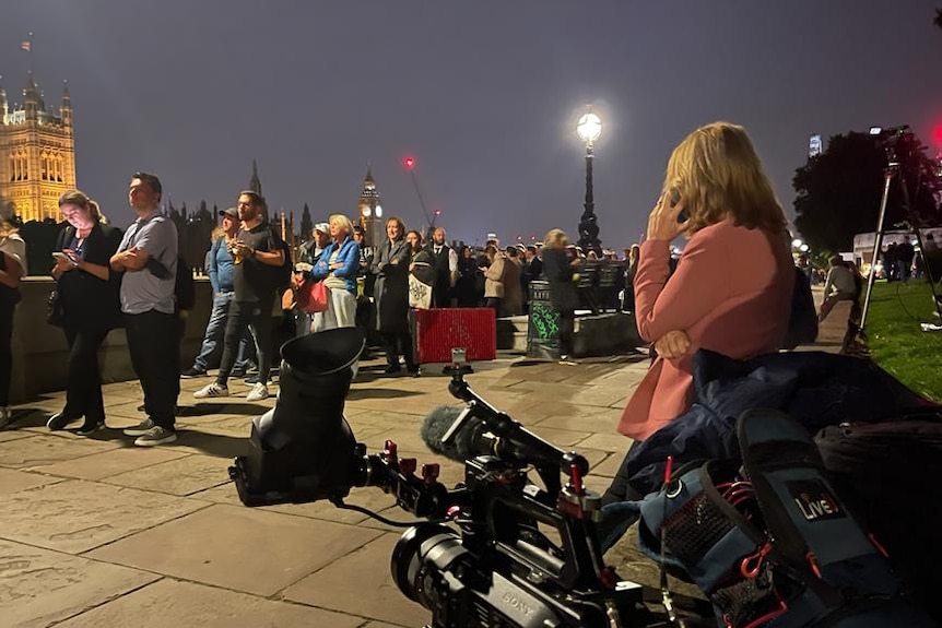 Night shot of journalist and TV camera pointed at queues of people on London street.