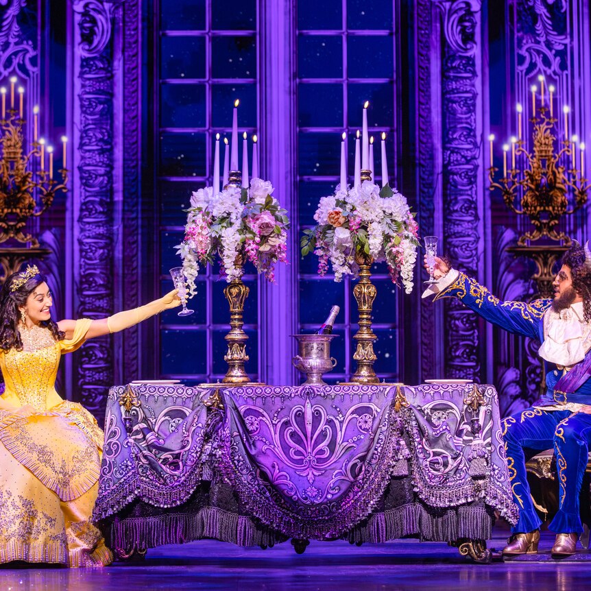 Stage production of beauty and the beast with Belle in a yellow dress sitting at one end of a grand table with beast opposite