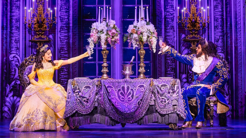 Stage production of beauty and the beast with Belle in a yellow dress sitting at one end of a grand table with beast opposite