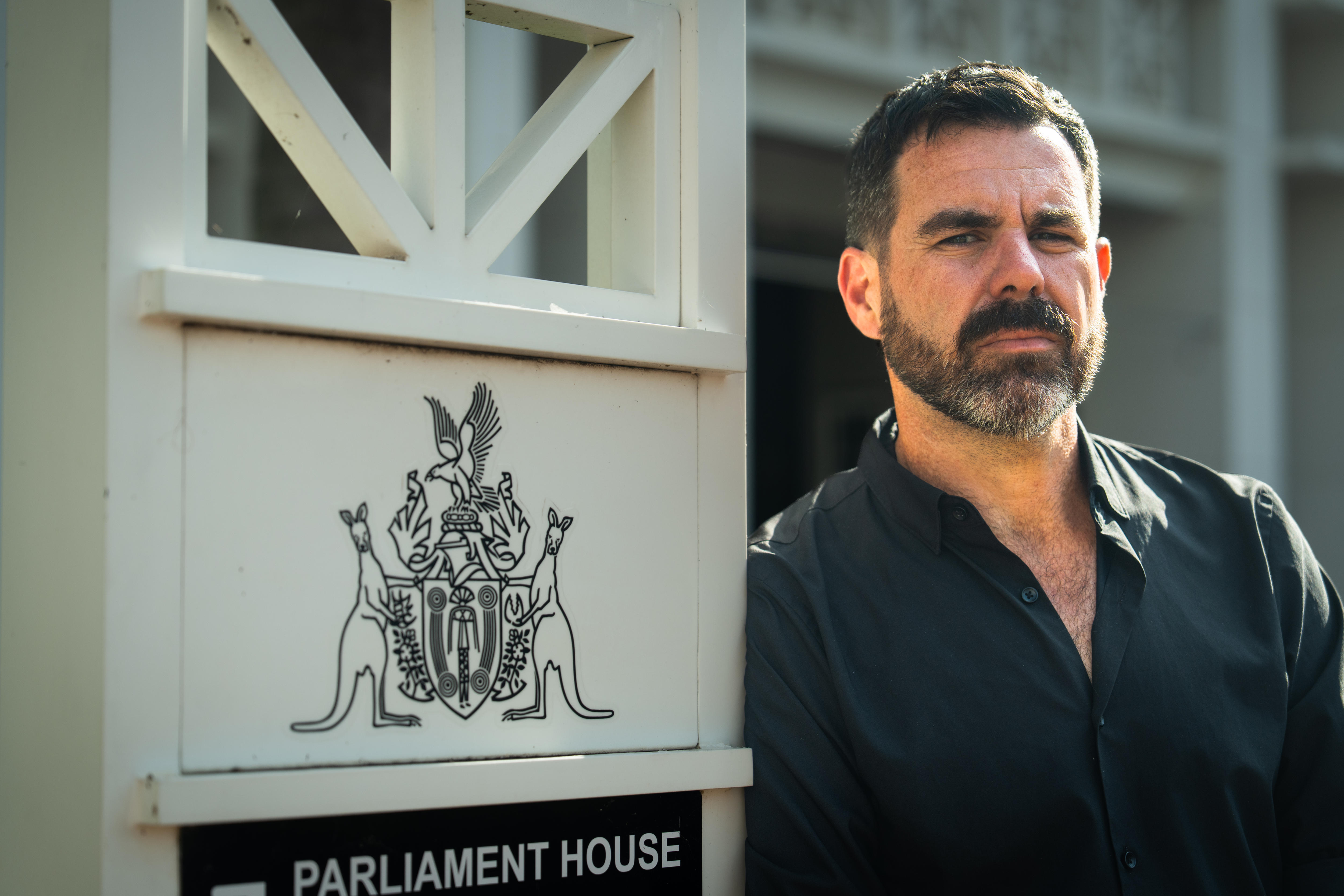 Man with dark brown short cut hair and brown, graying beard, standing and leaning on an NT parliament sign, serious expression.