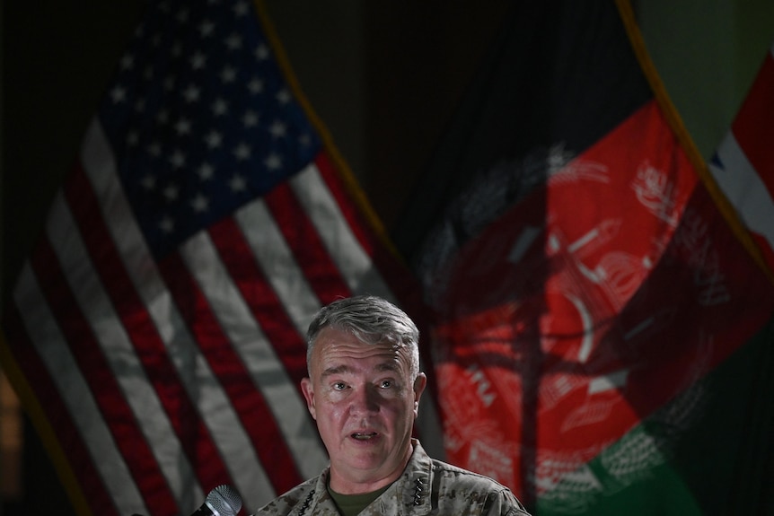 A military general in fatigues speaks in front of US and Afghan flags