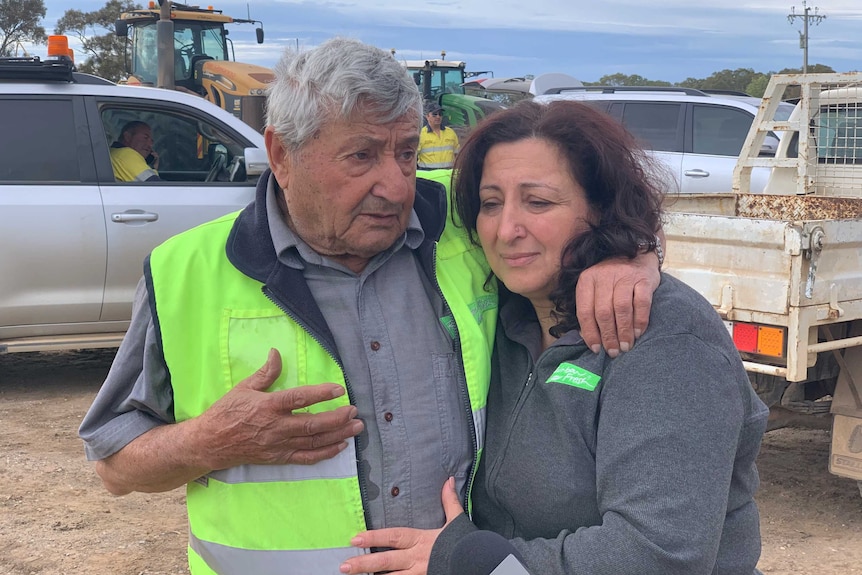 An elderly man and woman hug with cars and tractors in the background.