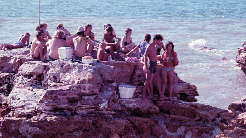 An archival, faded colour photo of a group of people on a rock off the Darwin coast.