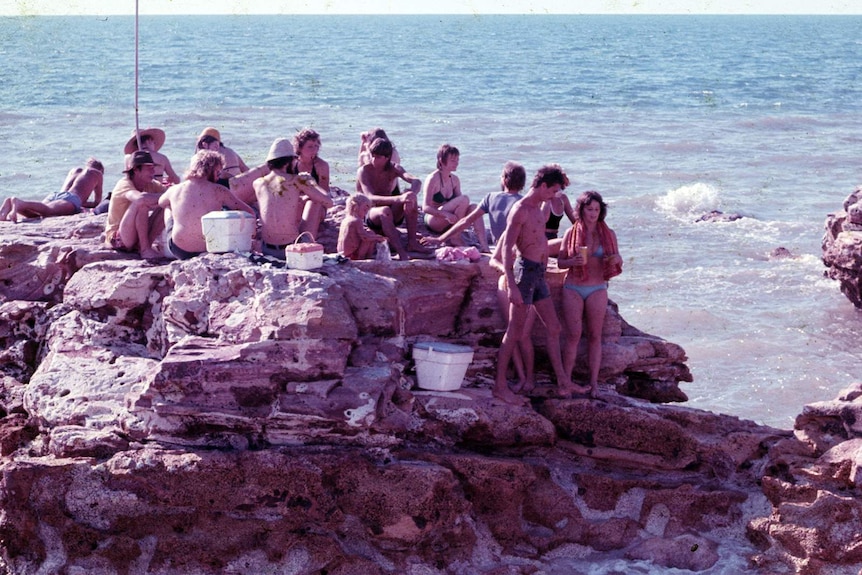 An archival, faded colour photo of a group of people on a rock off the Darwin coast.